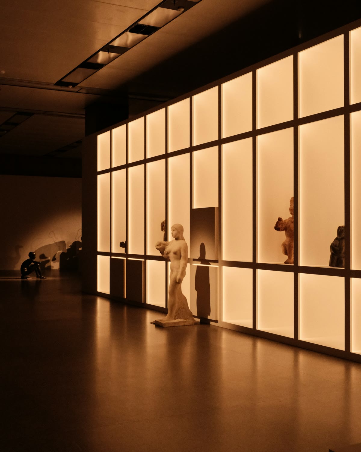 Sculptural forms on plinths in a warmly lit gallery, low ambient wash and long shadows